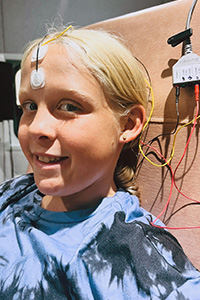 A young girl sits in a chair connected to monitoring equipment as part of an audiology assessment.