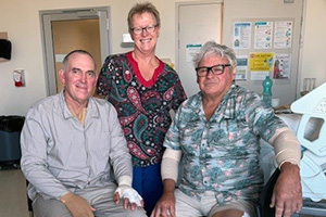 A female clinical nurse consultant stands between two older men who are seated. The man on the left has a bandage on his left hand.