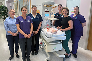 Six female Fiona Stanley Hospital maternity health professionals stand beside a mannuequin of a baby laying in a crib.