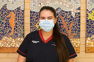A female nurse wearing a surgical mask stands in front of a tiled mosaic.