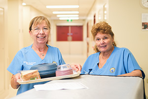 Two women wearing Fremantle Hospital Patient Support Services uniforms stand with a tray of food at a trolley
