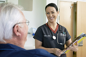 A female clinical nurse speaks to an older male patient seated in a bed