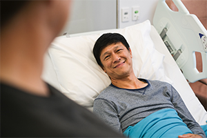 A man lies in a hospital bed smiling at a person standing beside him.