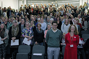 A crowd of 150 people standing together in the centre of a lecture theatre
