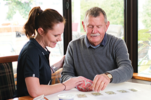 A female therapist sits with a male patient at a dining table in a home setting.