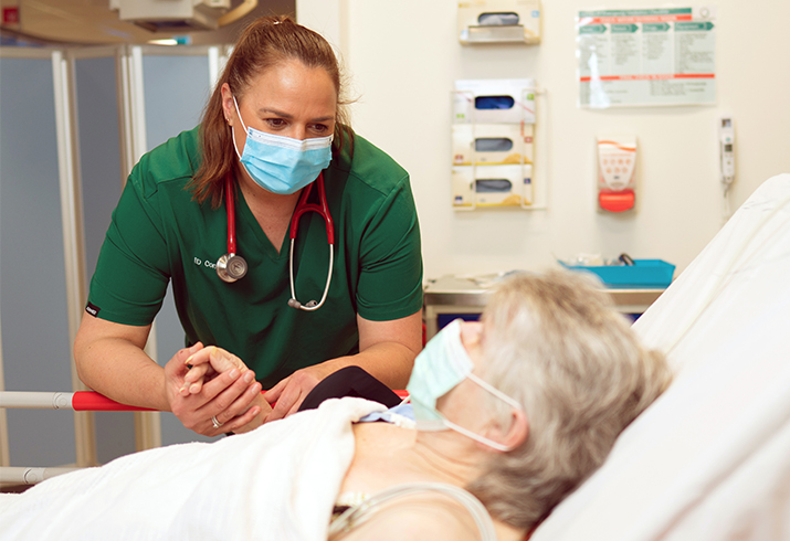 A female doctor holds the hand of an older woman in a hospital bed.