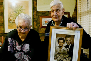 An older woman and man stand beside each other. The man holds a wedding photo of him and the woman.
