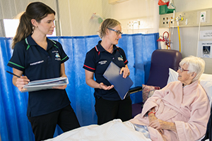 Two female allied health professionals stand beside the bedside of a female patient