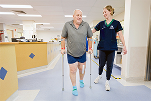 A female physiotherapist stands beside a male patient who is walking with elbow crutches in a hospital corridor