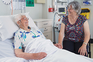 A smiling woman sits at the bedside of an older woman in a hospital bed