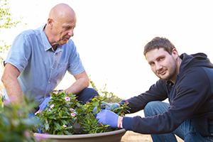 An older male health professional watches a younger man potting a plant in a large pot.