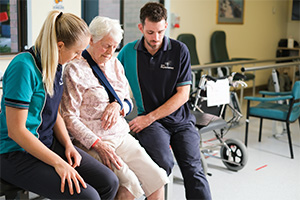 A male and female physiotherapist sit with an older woman in a rehabilitation gym.