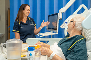 A female dietitian demonstrates to an elderly male patient how to order a hospital meal using an online system. A tray of food is in front of the man who is sitting in bed.