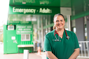 A female doctor stands outside a building. Over her shoulder is a sign that reads Emergency – Adults