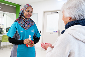 A female hospital volunteer wearing a headscarf offers some hand sanitiser to an other woman who is holding her hands in front in her.