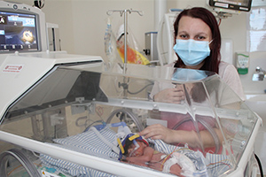 A young woman stands beside a small neonatal baby who is laying in a humidicrib