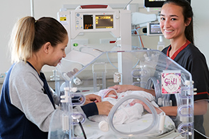 A young woman and a female nurse stand either side of a small neonatal baby who is laying inside a humidicrib.