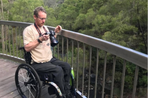 Clint sitting in a wheelchair on a bridge, looking at a photo he took on his camera of the view.