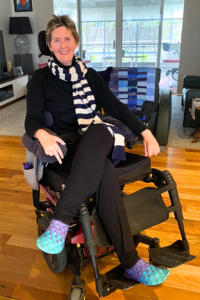 Kerry sitting in her wheelchair, cross legged with a scarf around her neck, smiling. 