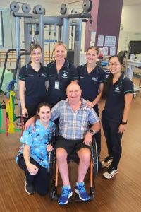 Kym pictured front sitting in wheelchair, alongside five female physiotherapists in the gym.