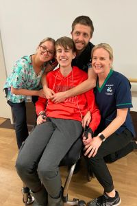 Lawson pictured middle sitting in a wheelchair wearing a bright red jumper, smiling alongside three staff on the ward.