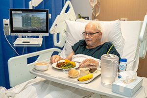 A tray of food is in front of an elderly male patient who is sitting in bed.