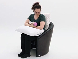 A woman holding a baby sits in a high backed chair. Her lower back is supported by a cushion and a pillow is underneath her arms to bring the baby closer for feeding