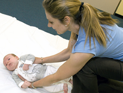 A woman bends over to pick up a baby laying on its back on the floor. The woman picks the baby up by rolling it gently side to side to place to place her hands under the baby’s armpits.