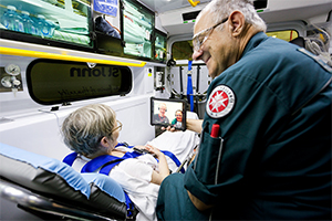 A female patient laying in an ambulance and a male paramedic beside talk to people via a digital tabled device