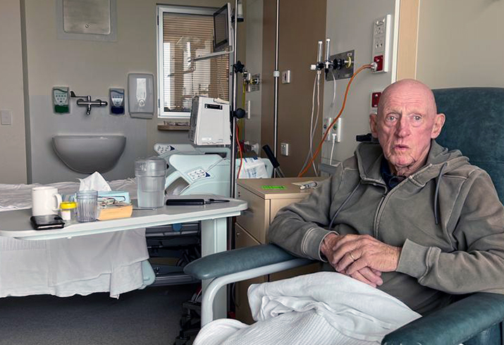 Bill Hazel pictured to the right, sitting in a chair in his hospital room. 