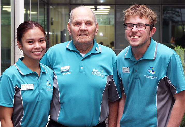 Three volunteers - one female and two males, wearing blue volunteer shirts, standing together smiling.  