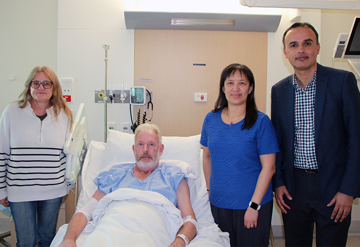 An older man lays in an hospital bed. A woman stands to his left, and another woman and a man stand on his right