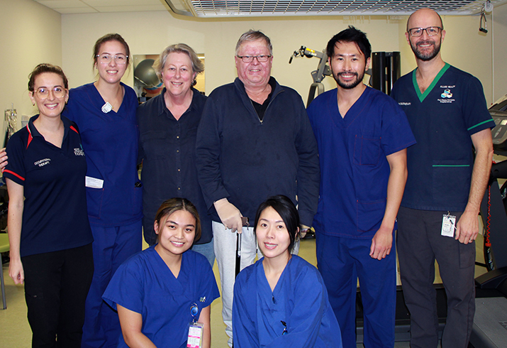 Male patient and his wife stand in a hospital rehabilitation gym surrounded by clinical staff, smiling.