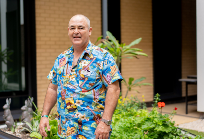 Man stands smiling in front of building and garden in a bright-coloured shirt.