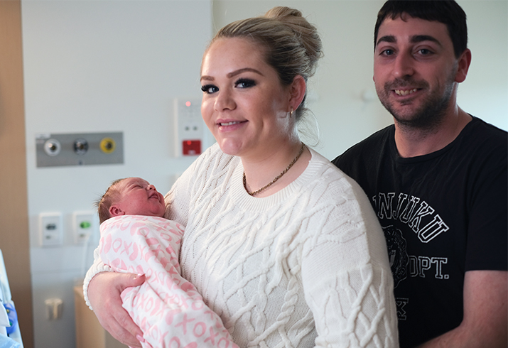 A young woman holds a newborn baby wrapped in a pick blanket. A young man stands behind her.