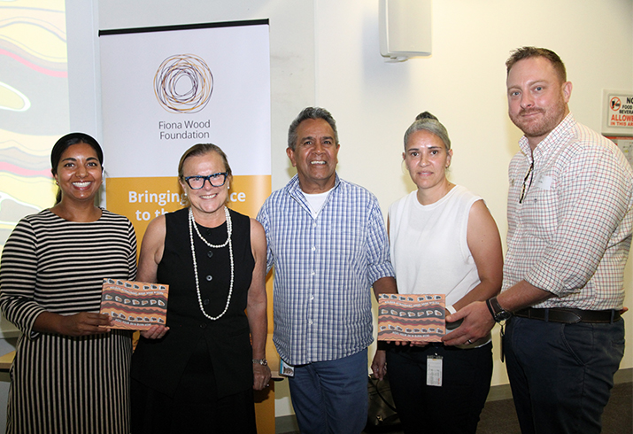 A group of five Aboriginal and non-Aboriginal men and women stand together. Two people are holding a small electronic tablet which features Aboriginal artwork on the cover.