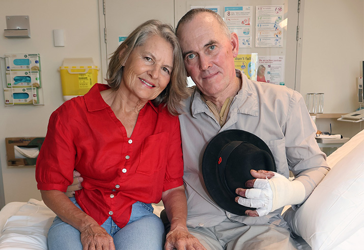 A woman and man sit beside each other on a hospital bed. The man's left hand is bandaged and holds a hat.