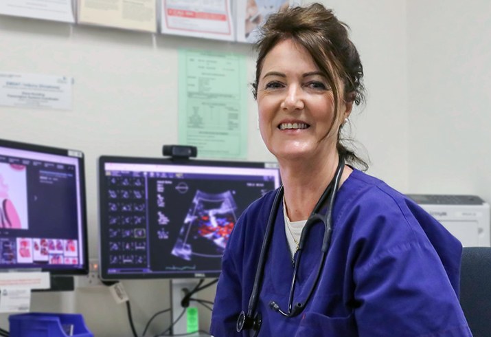 A female health professional sits at a desk. Medical images can be seen on two computer monitors behind her.