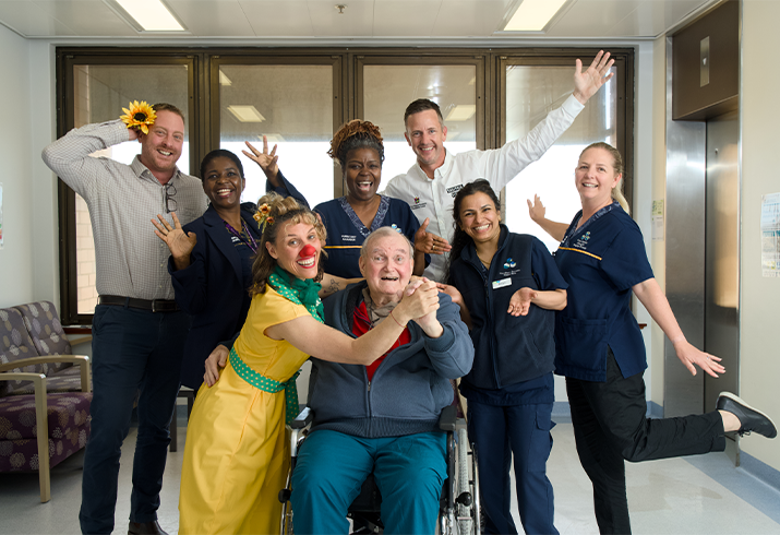 78-year-old Ken Westgate sitting in a wheelchair dancing with a Laughter Care Specialist who is wearing a bright yellow dress, surrounded by six cheerful staff.