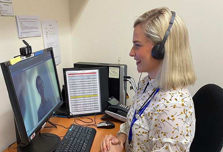 A female clinician wearing headphones sits in front of a computer monitor during a virtual telehealth appointment. A patient can be see on the montior.