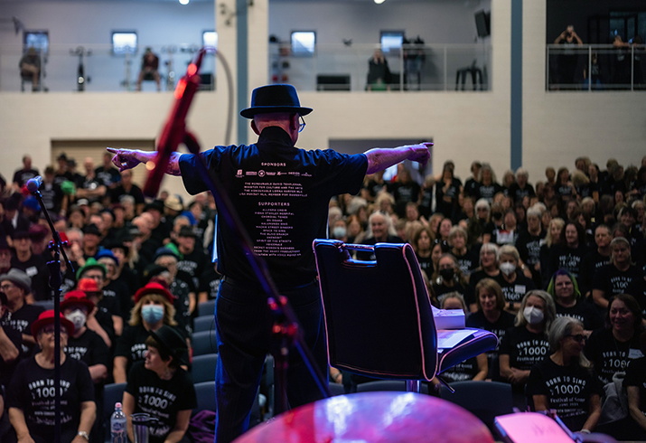 A man with his back to the camera stands on a stage conducting a choir