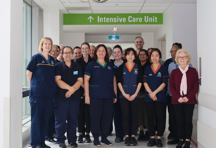 Twenty staff members from the Fiona Stanley Hospital Intensive Care Unit standing together in a hallway, under a green wayfinding sign that says "Intensive Care Unit".