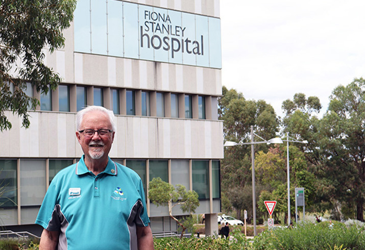 Paul Hammond stands in front of Fiona Stanley Hospital wearing a Fiona Stanley Fremantle Hospitals Group volunteer shirt 