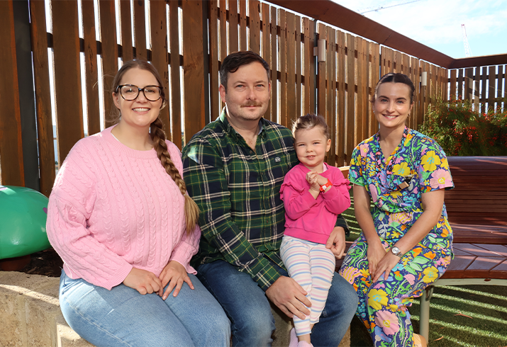 Matilda wearing a pink jumper pictured middle sitting on her dad's lap, with mum and nurse either side.