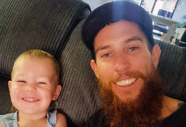 A young child sits on a couch next to a young man with a beard and mustache.